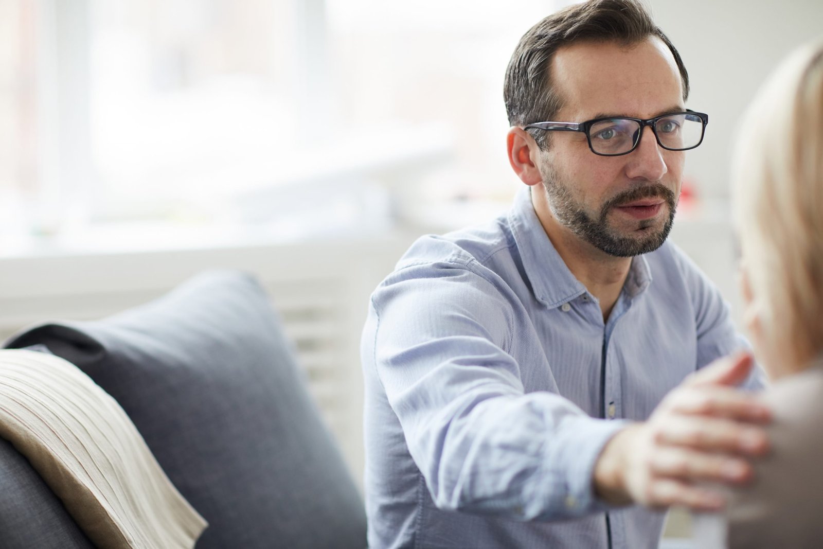 Mature counselor in shirt and eyeglasses touching shoulder of patient while comforting her during session