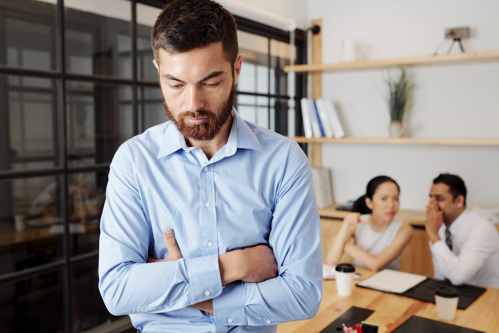 Upset young businessman standing in closed pose, colleagues are gossiping about him in background