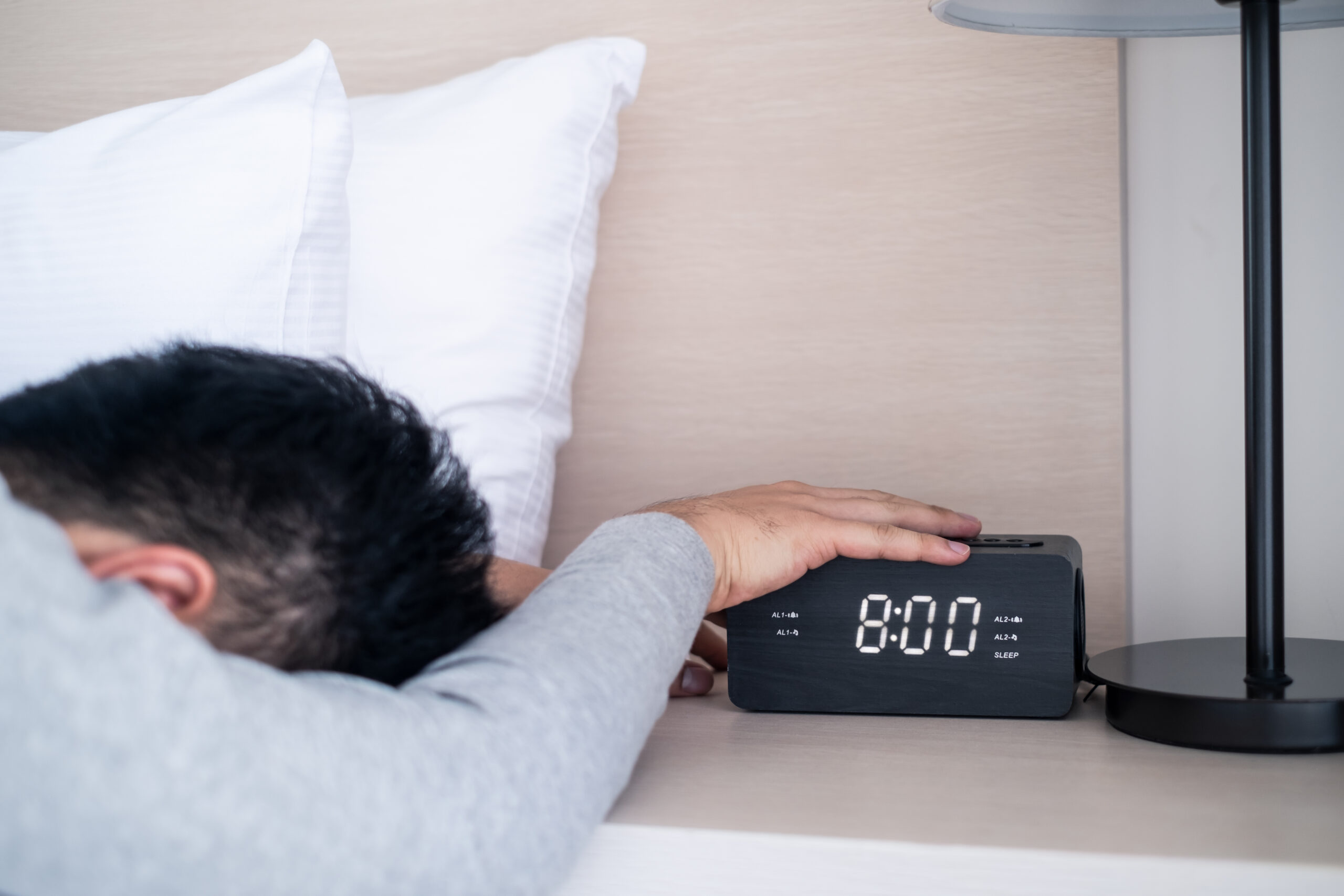 Lazy male office worker sleeping on bed in bedroom, stopping alarm clock in the morning on weekday. Man put hand on digital clock button to silent the voice and feeling bore to wake up, feels sleepy.