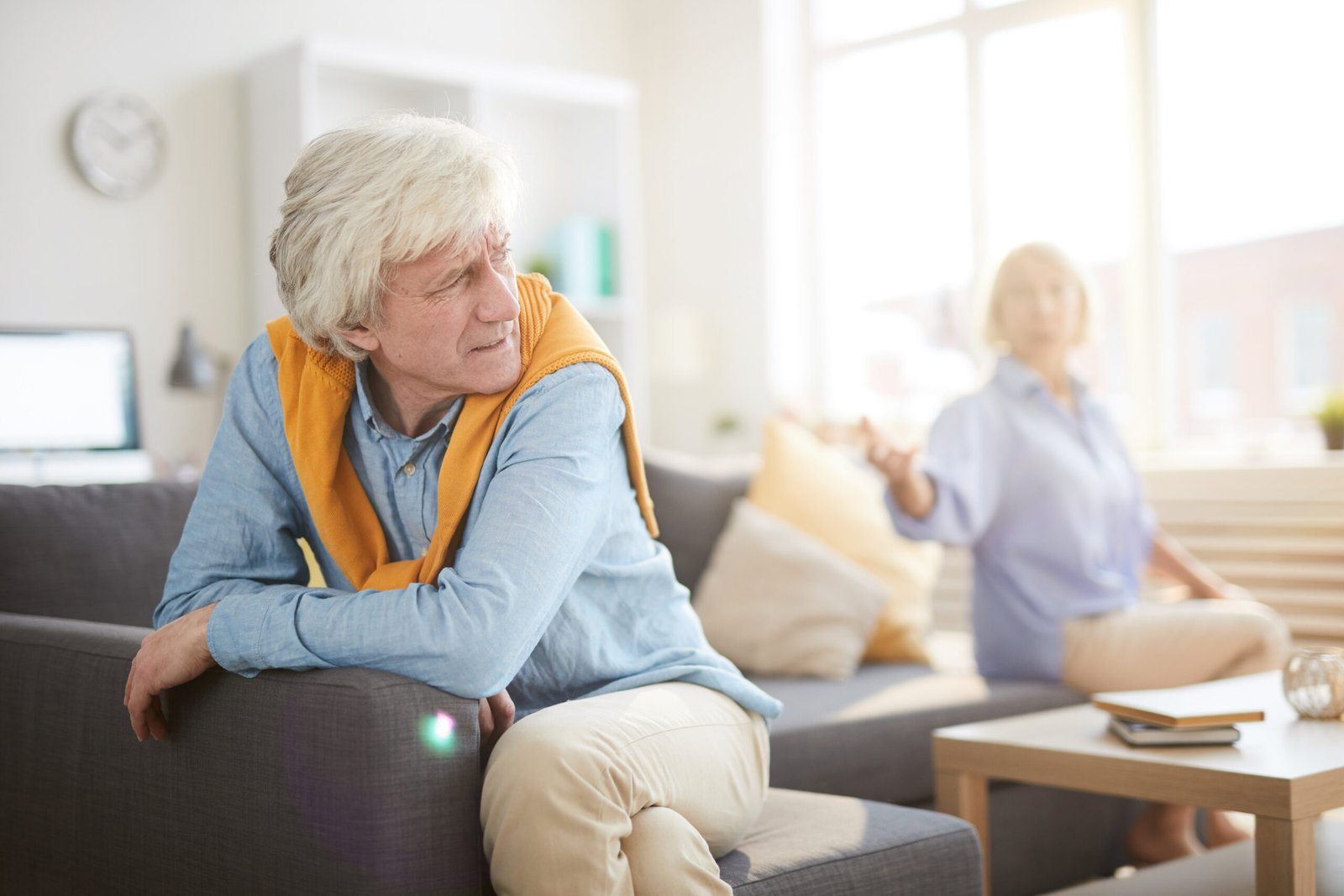 Portrait of senior couple fighting sitting on opposite sides of sofa at home, focus on angry husband in foreground, copy space
